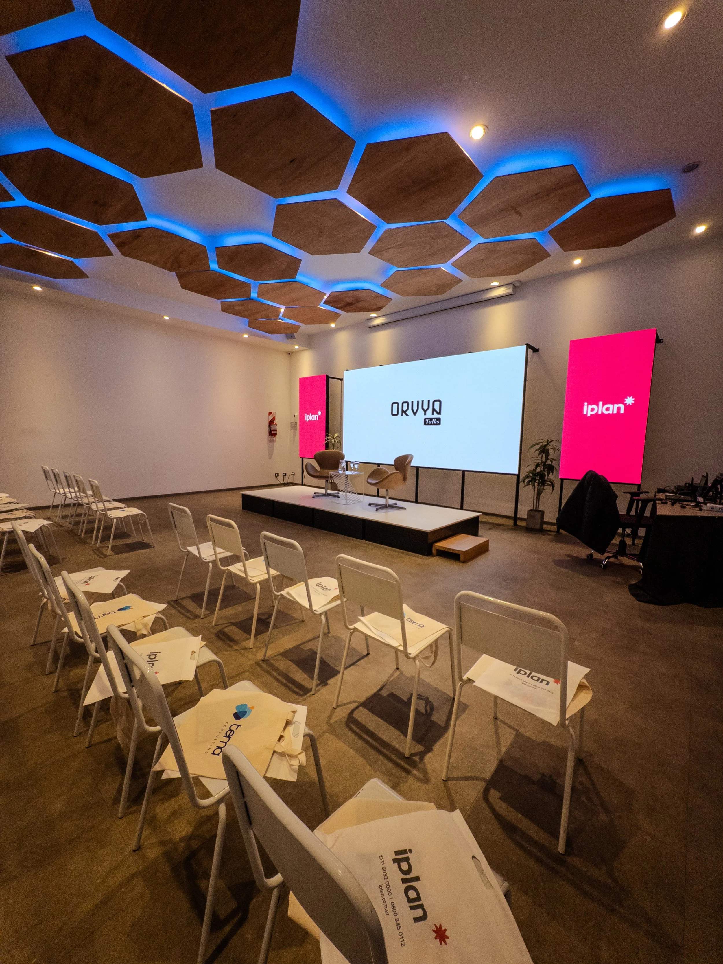 Sala de conferencias con pantalla y sillas, decorada con paneles hexagonales en el techo y luces azules.