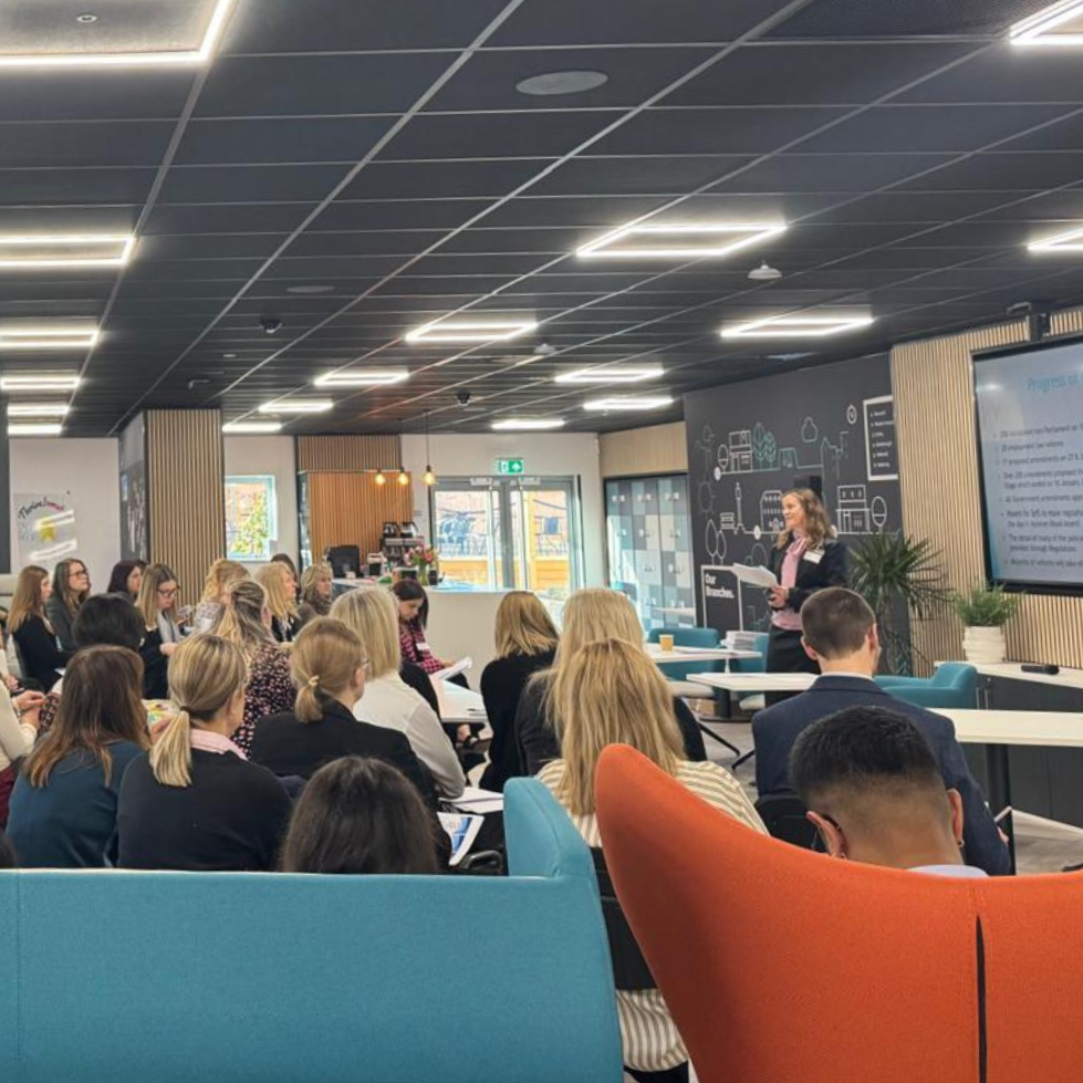 A woman gives a presentation to a group of people in a modern conference room with a large screen and audience seated in colorful chairs.