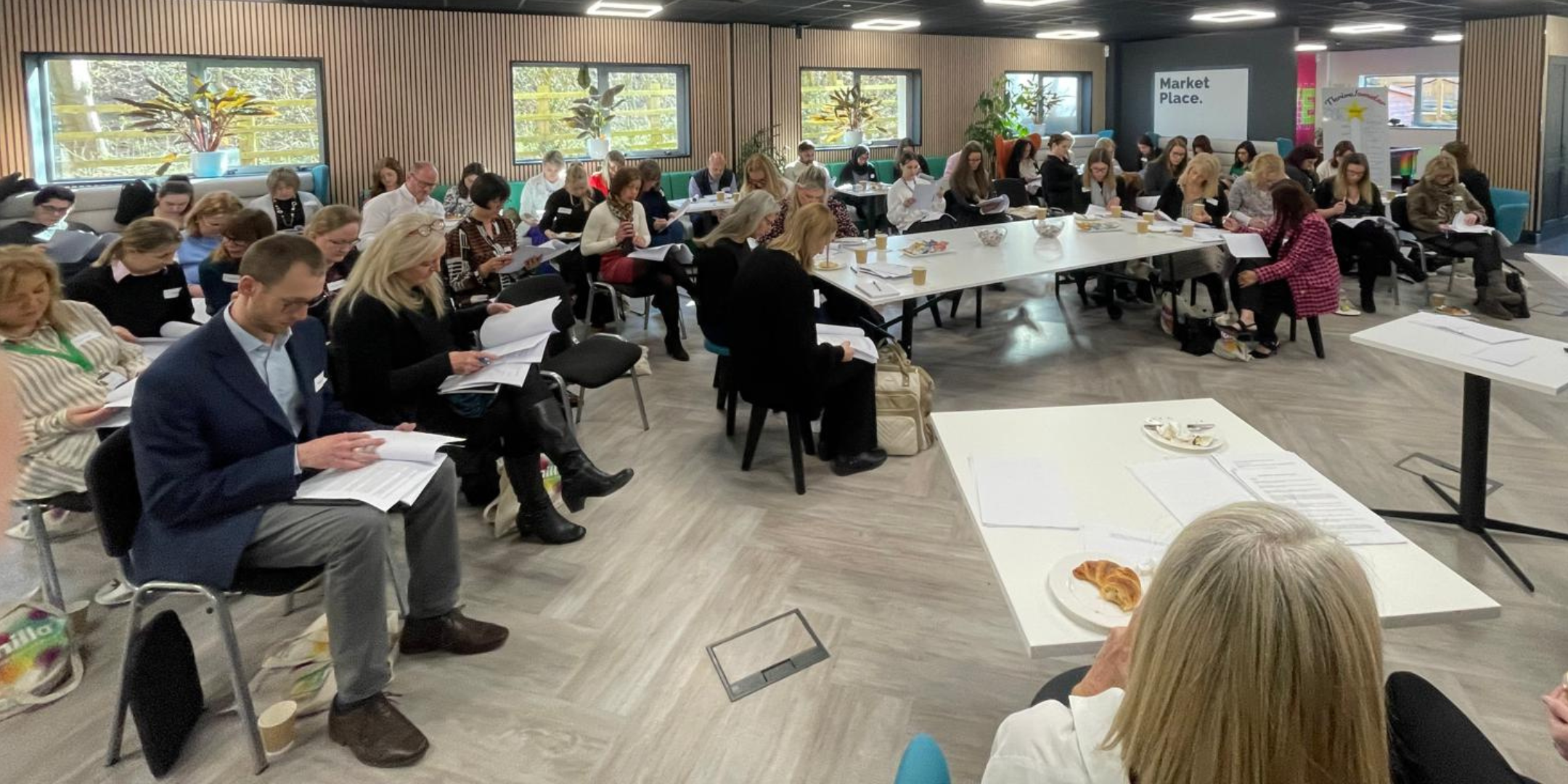 A large group of people attending a conference or seminar in a modern room with wood wall panels, large windows, and plants. They are seated at tables or chairs, reading or listening. There are some plates with snacks, paper cups, and notebooks on the tables. The overall atmosphere is professional and attentive.