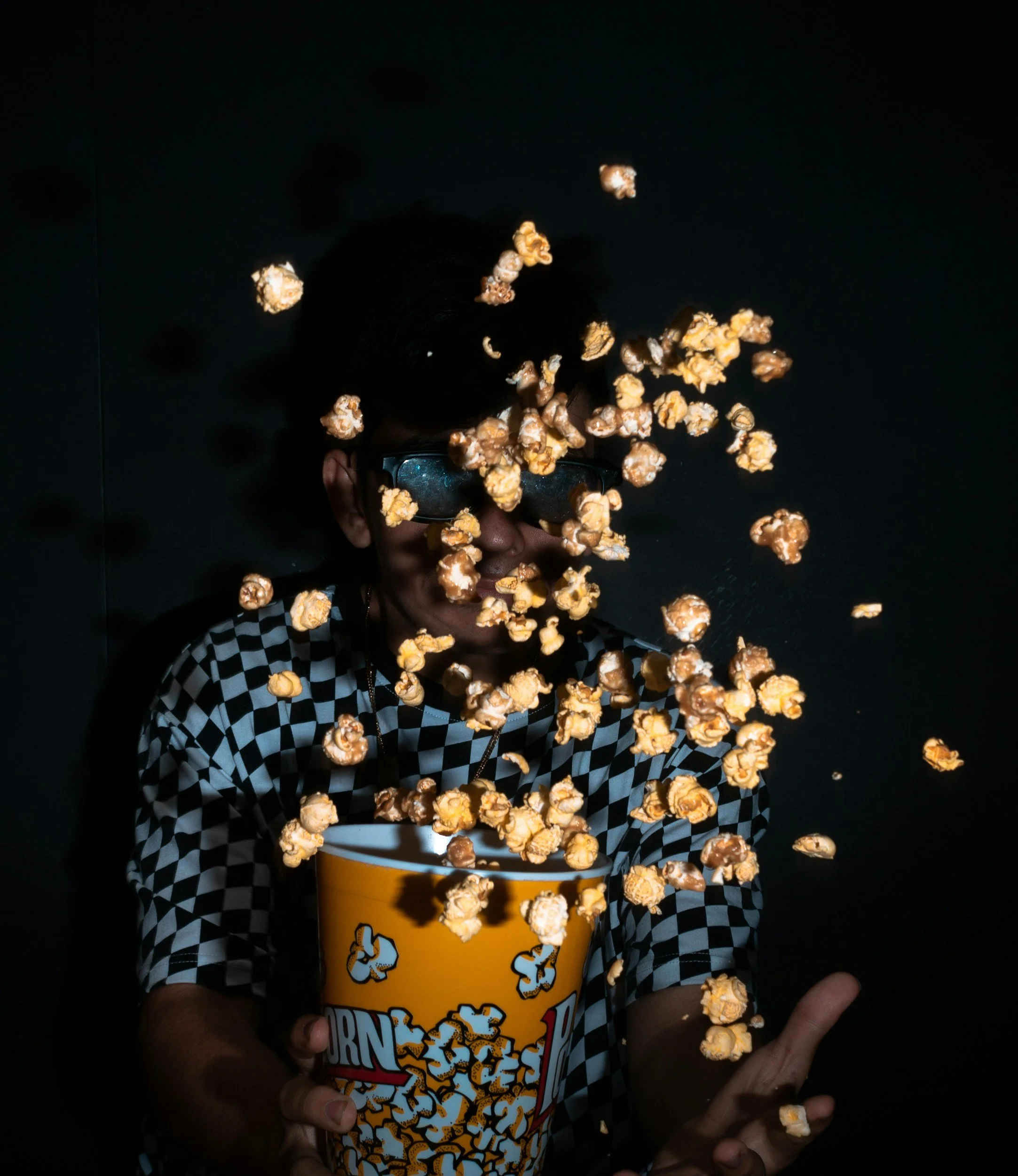 Person wearing black glasses and a black-and-white checkered shirt holding a large yellow popcorn bucket with popcorn flying in the air against a black background.