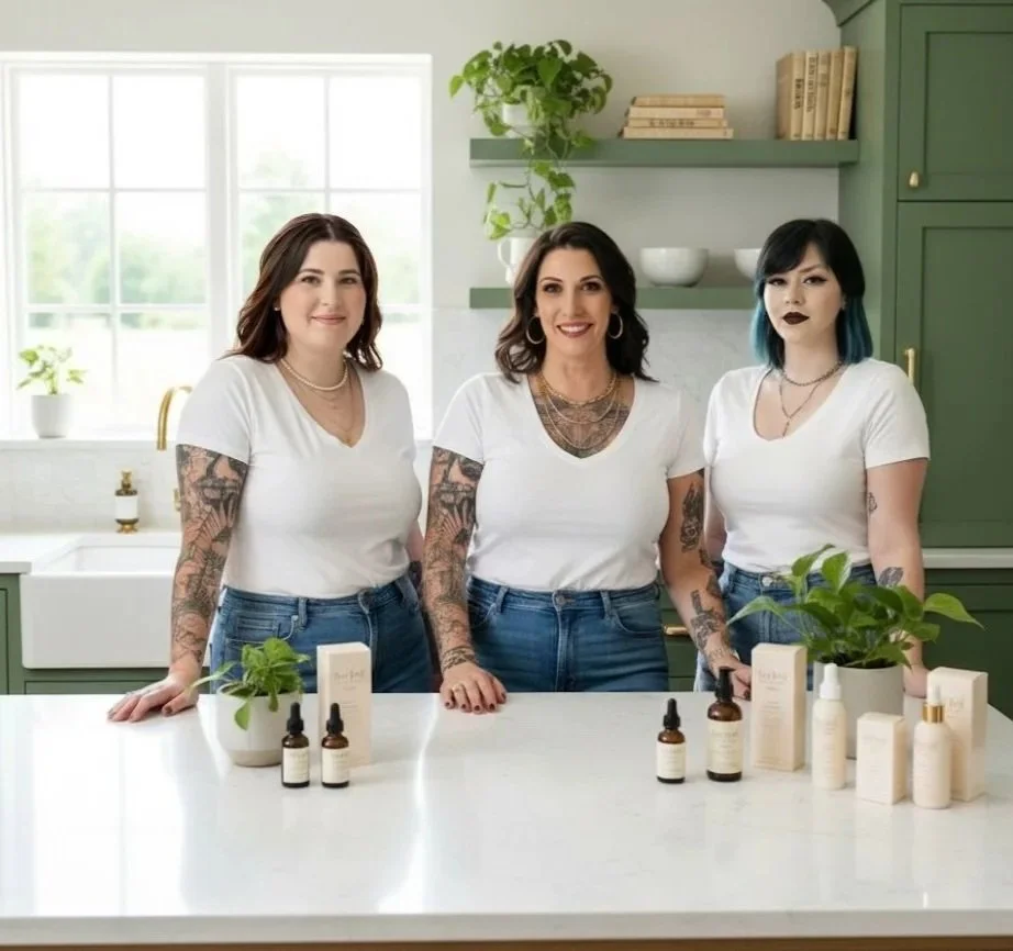 Three women with tattoos standing behind a kitchen counter with skincare products and plants, smiling at the camera.