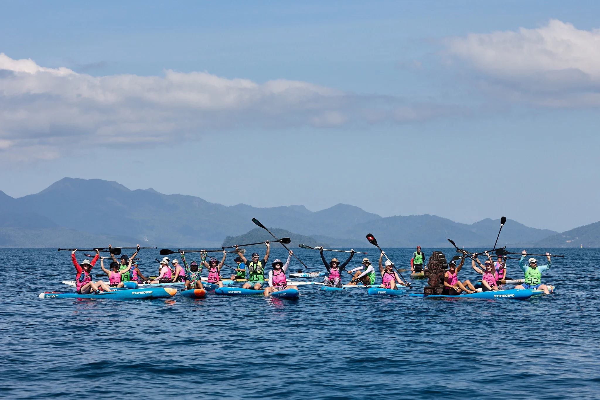 Group of people paddleboarding on the ocean during daytime, with mountains and cloudy sky in the background.