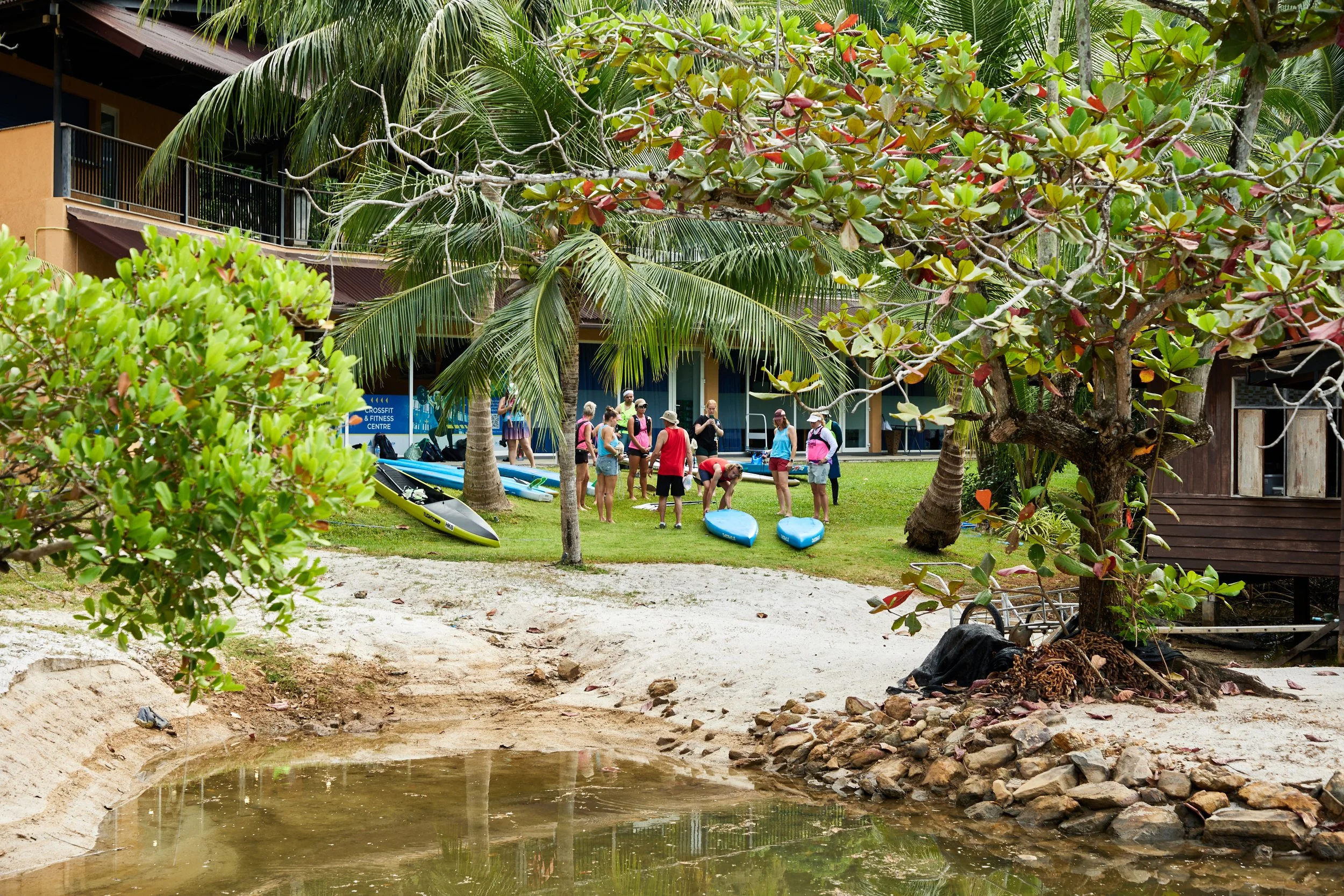 Group of people preparing for paddleboarding at a tropical resort with palm trees, a sandy area, a small pond, and a building in the background.