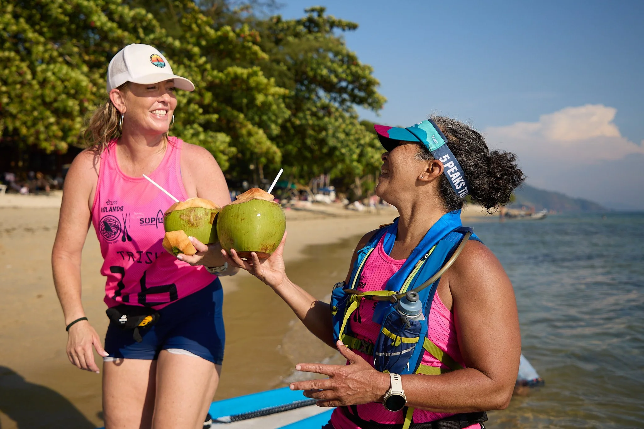 Two women in athletic gear on a beach exchanging coconuts with straws, smiling at each other, with trees and boats in the background.