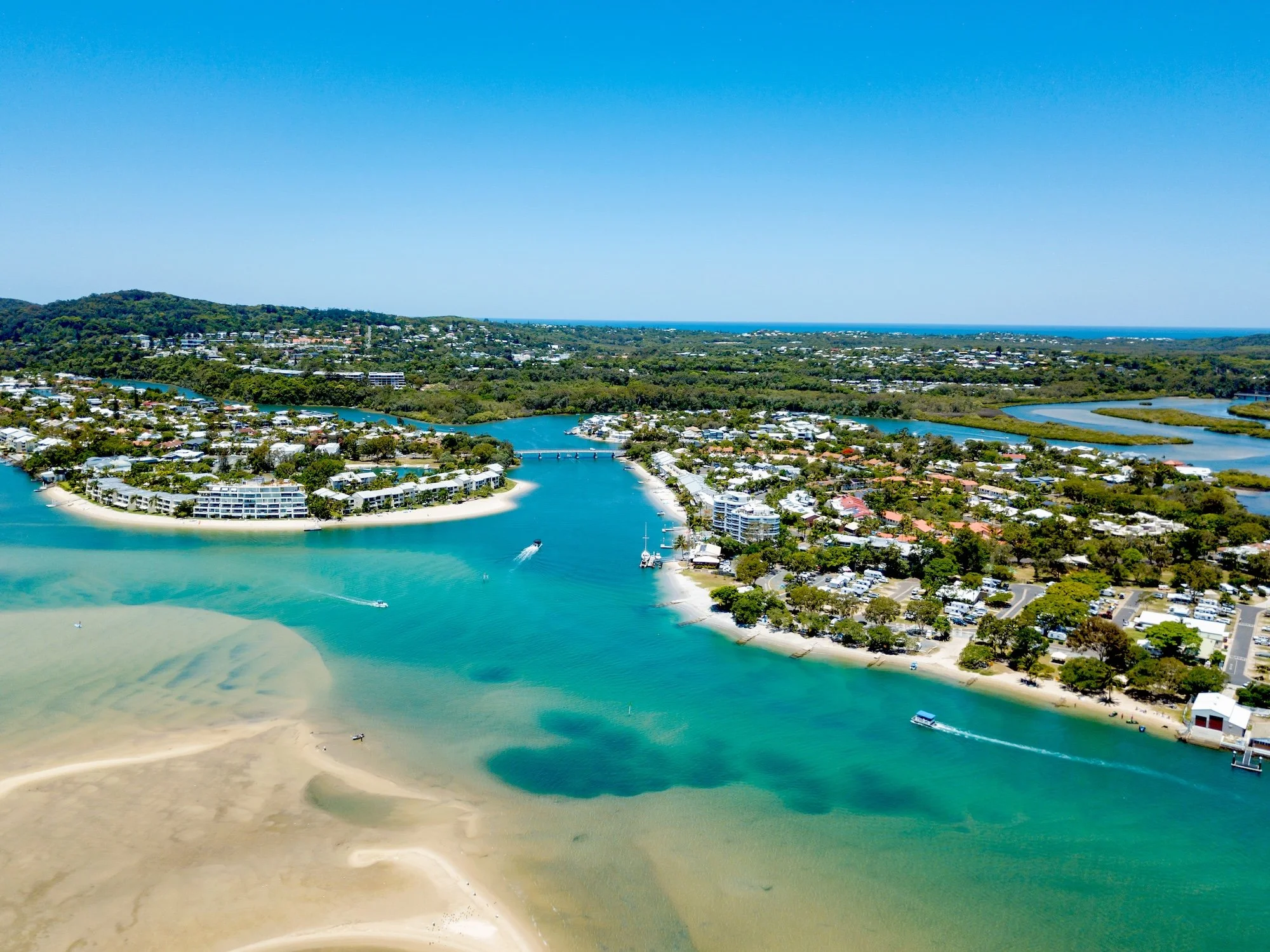 Aerial view of a coastal town with a marina, waterways, and green hills in the background under a clear blue sky.