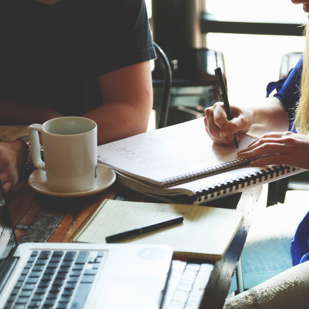 People are sitting at a table working on notebooks, with a laptop, a coffee mug, and a notepad visible.
