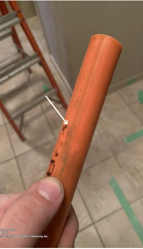 A person's hand holding an orange PVC pipe with a small black mark on it, in a room with tiled floor and exposed ladder.