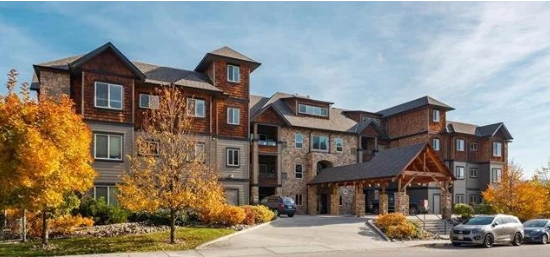 A multi-story residential building with a stone and wood exterior, surrounded by autumn-colored trees and parked cars under a partly cloudy sky.