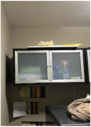 A silver and black cabinet mounted on a beige wall, with a yellow item and a cloth on top, and some books and a box on the shelves below with Plumbingtrack installed above at the wall to ceiling transition