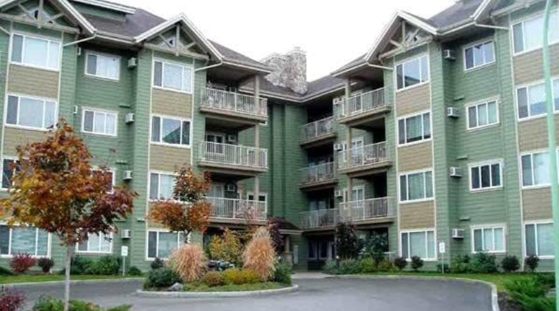 A multi-story apartment complex with green and beige exterior, featuring several balconies and windows, surrounded by trees and landscaped bushes.