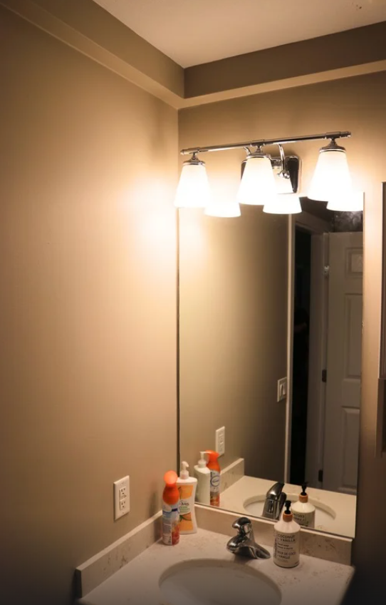 plumbing track installed and painted Bathroom vanity with a sink, soap dispenser, cleaning spray, and bottles, lit by four-light fixture, mirror reflecting a doorway.