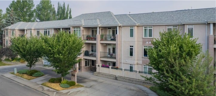 A three-story residential apartment building with balconies, trees, and a paved parking area in front.