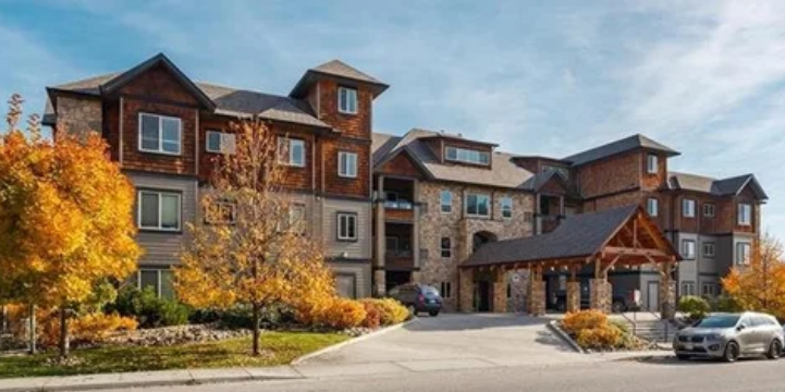A multi-story apartment complex with stone and wood exterior, surrounded by autumn-colored trees, with cars parked in front and a clear blue sky.