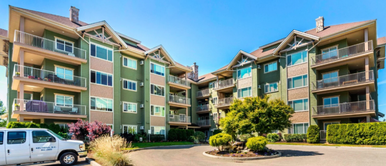 Exterior view of a multi-story apartment complex with balconies, surrounded by landscaping and a parking lot with a white van, under a clear blue sky.