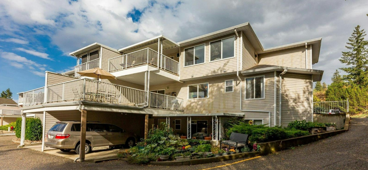 Multi-story beige residential building with balconies, a parking area underneath, and a garden with flowers in front.