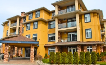 Yellow multi-story apartment building with balconies, brick accents, and a covered entrance with stone pillars, surrounded by shrubbery.