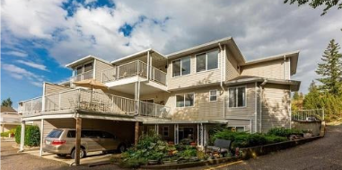 A multi-story residential building with balconies, surrounded by trees and a parking area with cars, under a partly cloudy sky.
