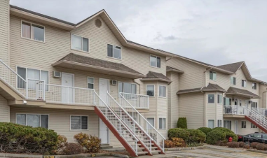 Multi-story beige apartment building with outdoor stairs, balconies, and landscaped bushes under an overcast sky.