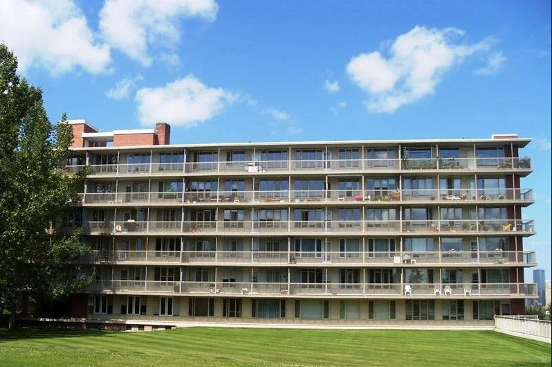 A mid-rise apartment building with multiple balconies, large windows, and a well-maintained grassy lawn in front, under a bright blue sky with scattered clouds.