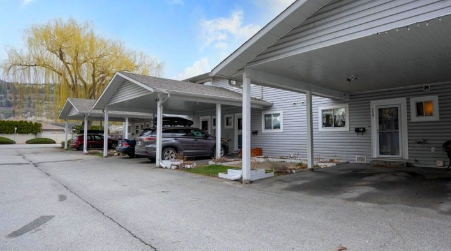 A covered parking area with several parked cars in front of a light gray building with white trim and windows.