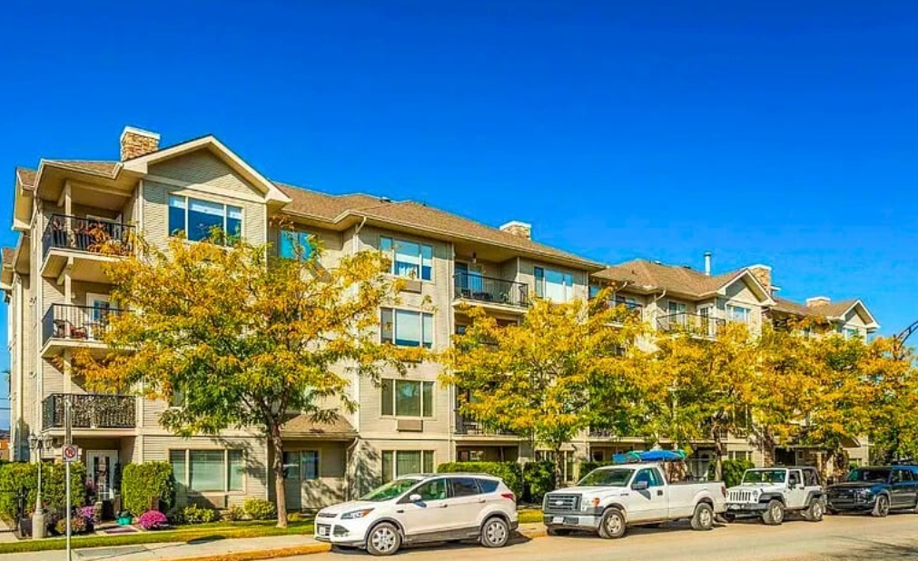 Multi-story apartment building with trees and parked cars in front, under a clear blue sky.