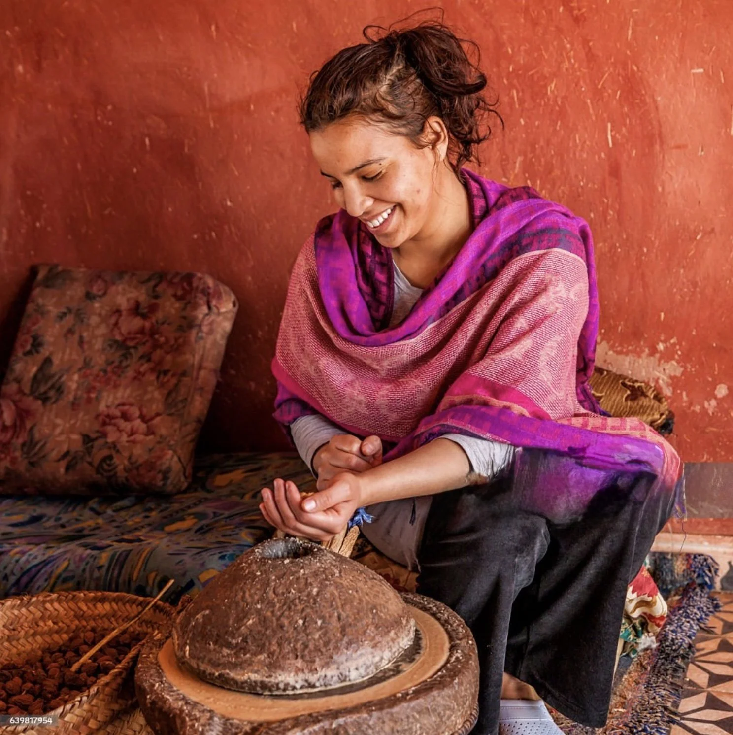 Une femme souriante, vêtue de vêtements colorés, utilisant un moulin à grain traditionnel dans une pièce à murs rouge/orangé.