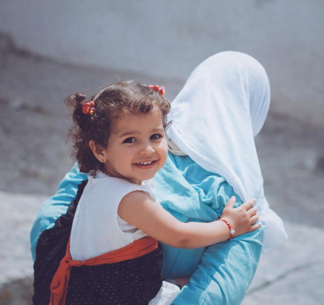 Une jeune fille souriante avec des cheveux bouclés, portant une chemise blanche avec une robe foncée à pois et une ceinture orange, en train de grimper ou de s'accrocher à une personne portant des vêtements bleus, devant un mur gris