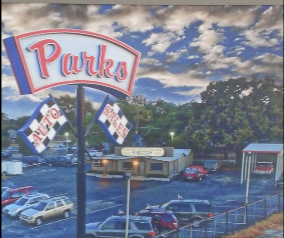 Outdoor parking lot with cars, a building, and a large sign reading 'Parks' with additional signs for 'Auto' and 'Sales', under a cloudy sky.