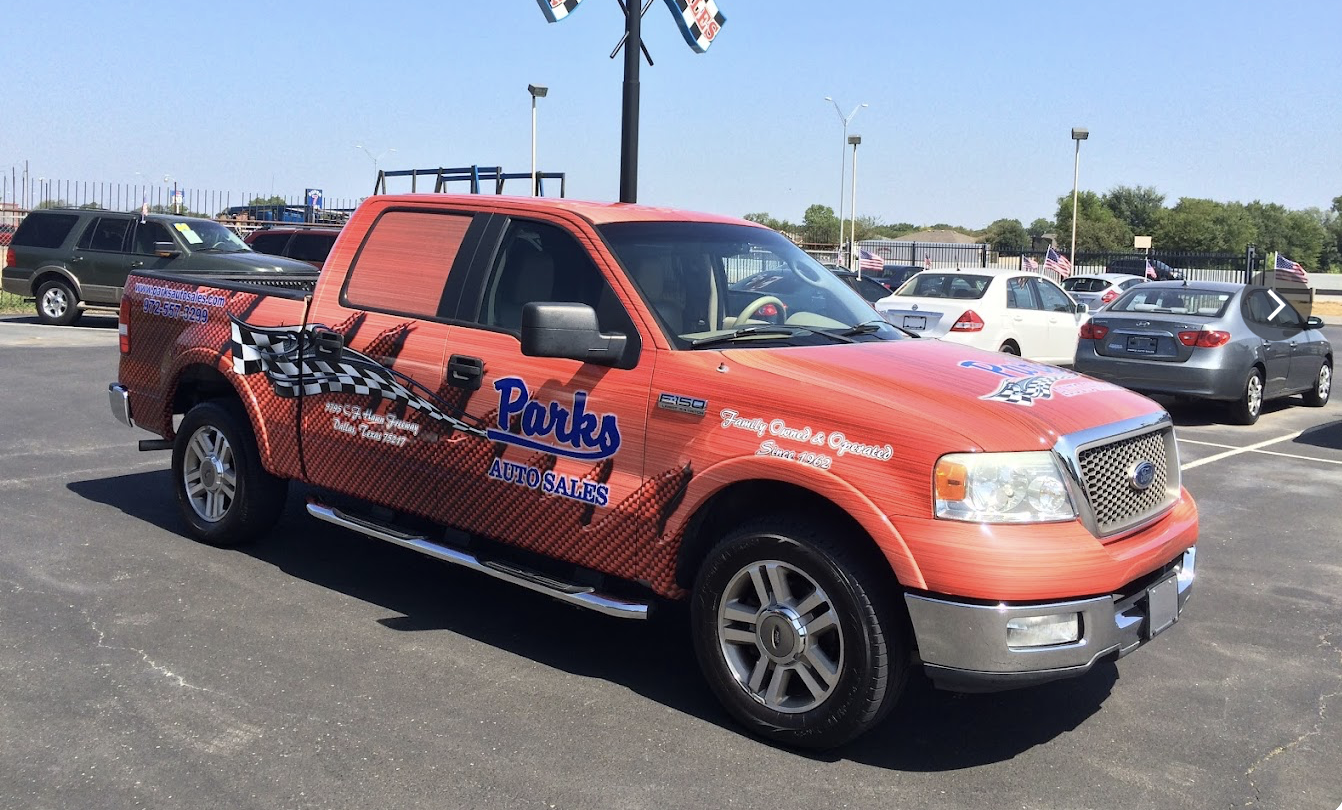 Red Ford F-150 truck with Parks Auto Sales advertisement parked in a car dealership lot, with other vehicles and American flags in the background.