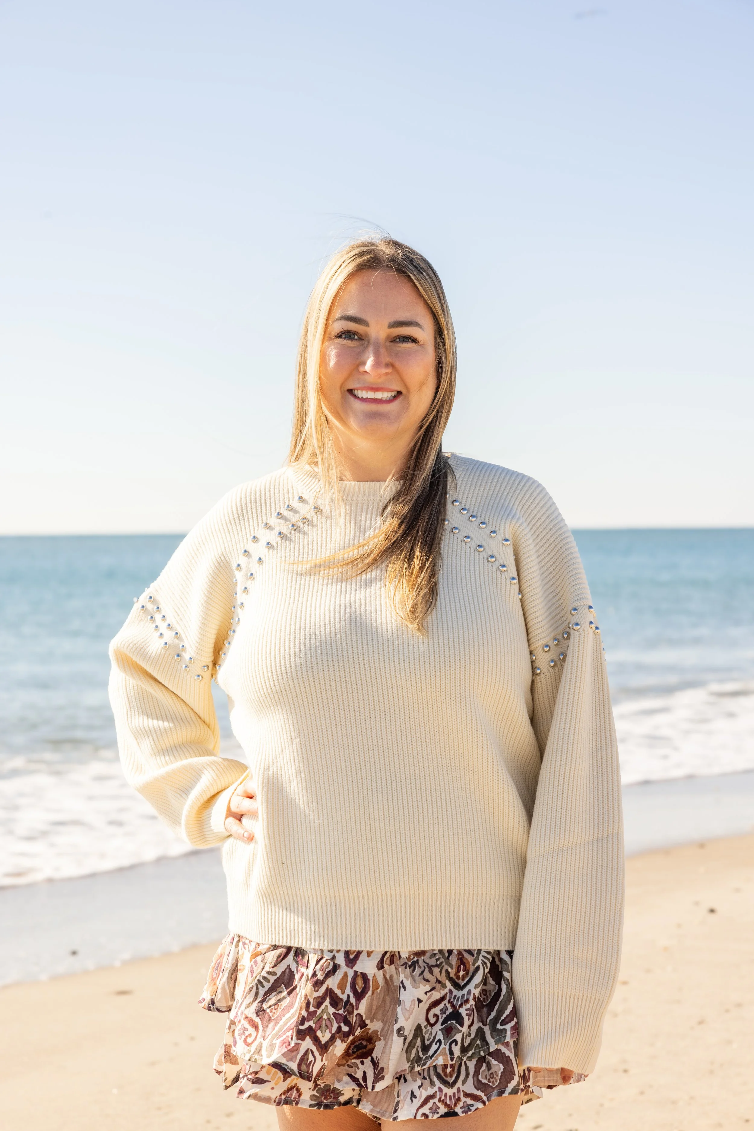 Smiling woman standing on a beach with the ocean in the background, wearing a cream sweater and patterned skirt.