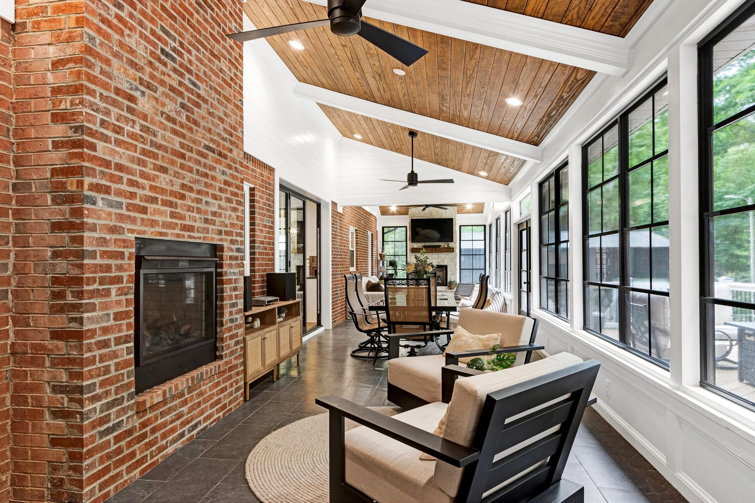 Sunroom with brick wall, wood-paneled ceiling, ceiling fans, large black-framed windows, living area with beige armchairs, a TV, and a fireplace.
