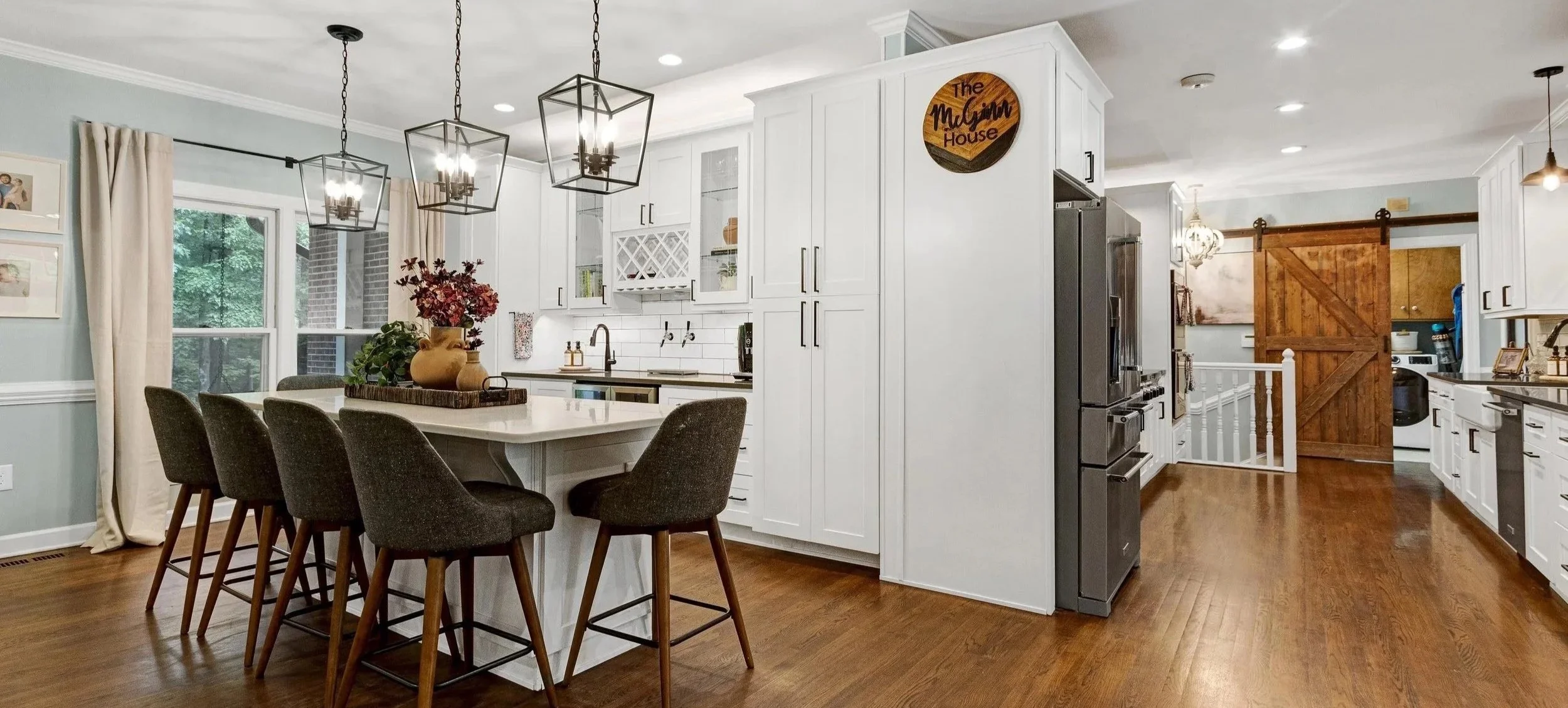 Modern kitchen with white cabinets, a center island with seating, and wooden flooring. There are pendant lights above the island, windows with curtains, and decorative items on the countertops. A sliding barn door leads to another room, and a round sign reads "The Mylgine House."