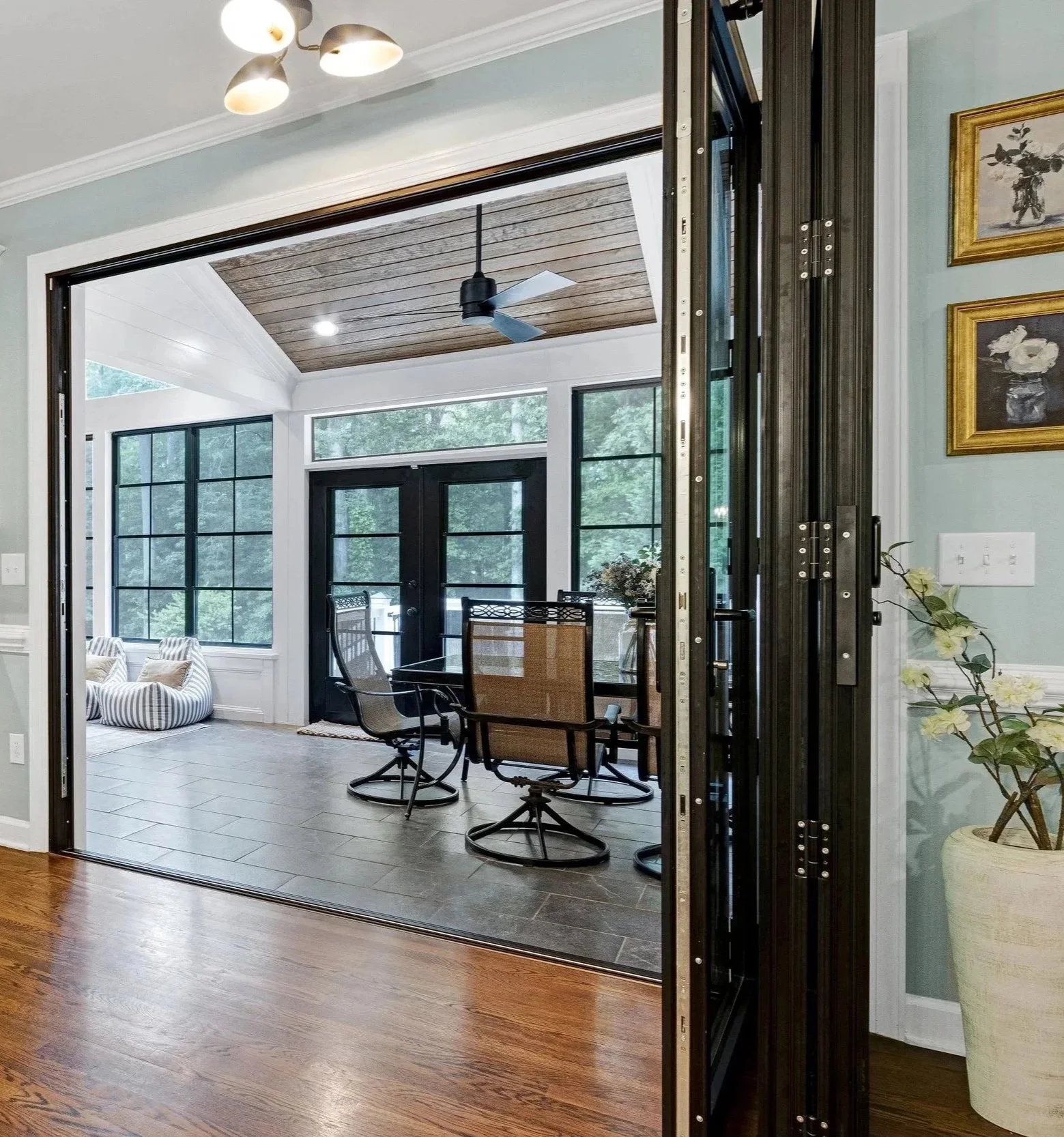 Interior view of a sunroom or enclosed porch with large windows, dark framed door, ceiling fan, and a dining table with chairs.