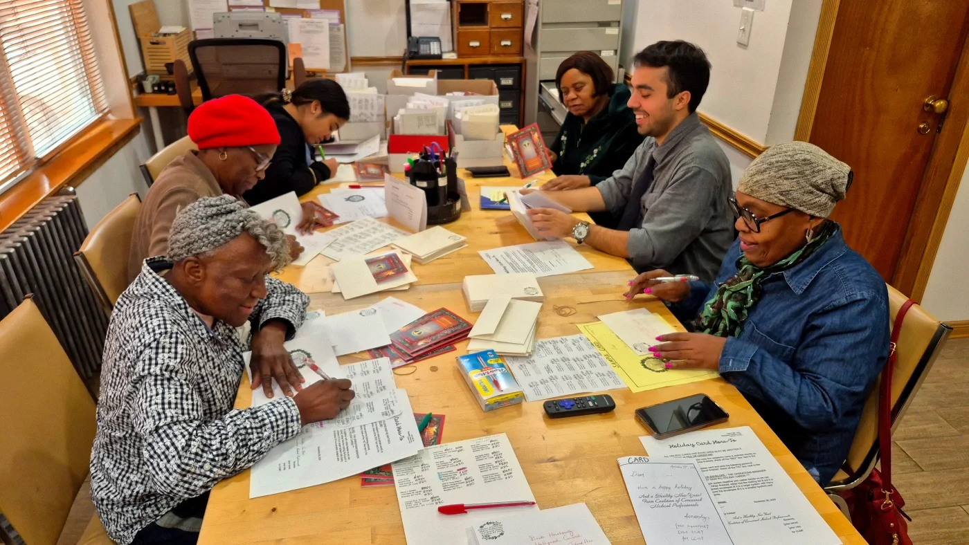 A group of six CCMp volunteers work on writing and mailing letters at a wood table in CCMP's office.
