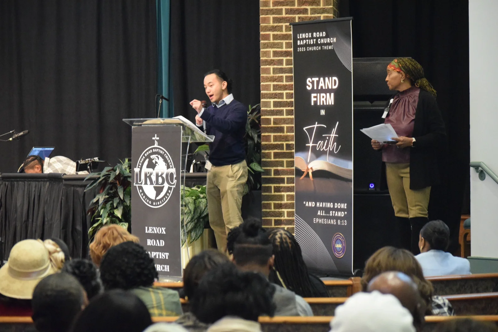 A volunteer addresses a crowd at a house of worship while a nurse volunteer accompanies him at the podium.