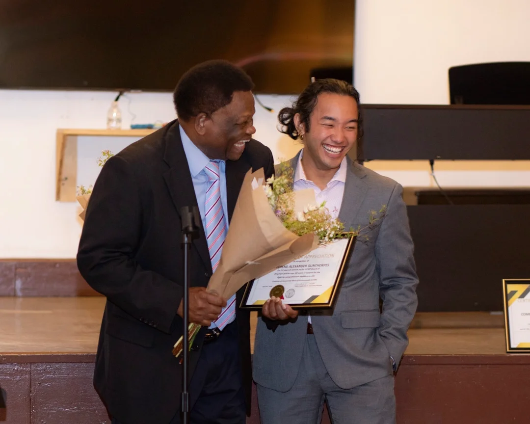 Two CCMP volunteers stand in front of a microphone, one holding an award and a bouquet of flowers. 