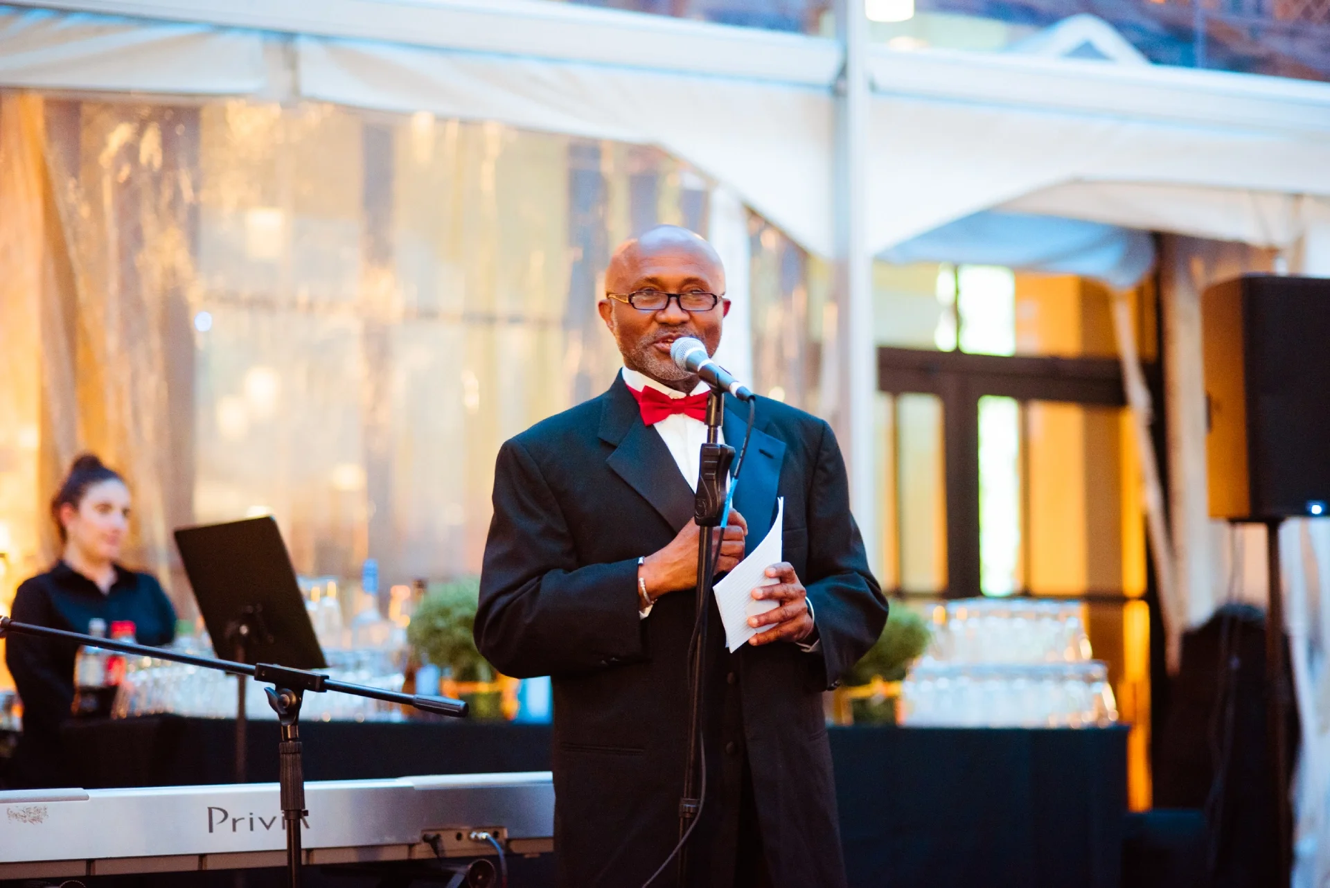 A volunteer wearing a black suit with a red bowtie speaks into a microphone and holding a small white paper. 