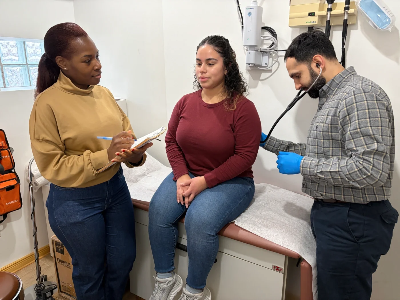 A volunteer doctor checks a benefit recipient's heart with a stethoscope  while a CCMP volunteer takes notes. 