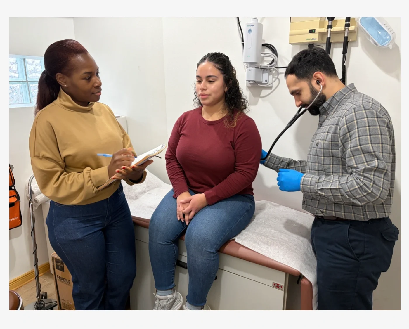 A volunteer doctor using a stethoscope to check someone's heart rate while a volunteer nurse and advocate takes notes.