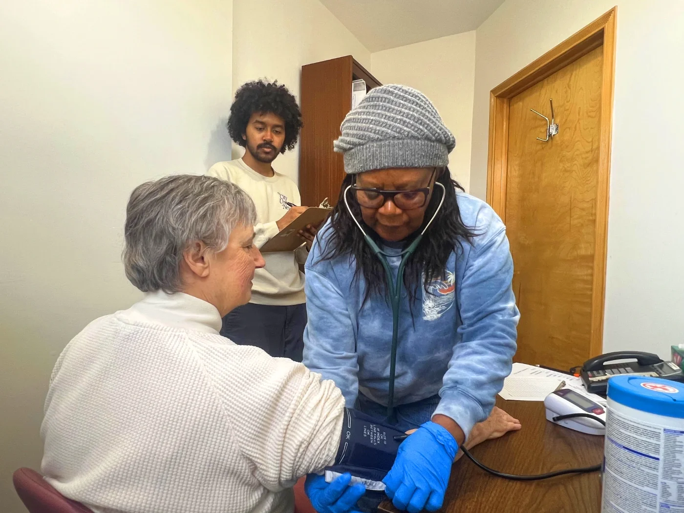 A volunteer nurse putting a blood pressure cuff on someone while an advocate takes notes.