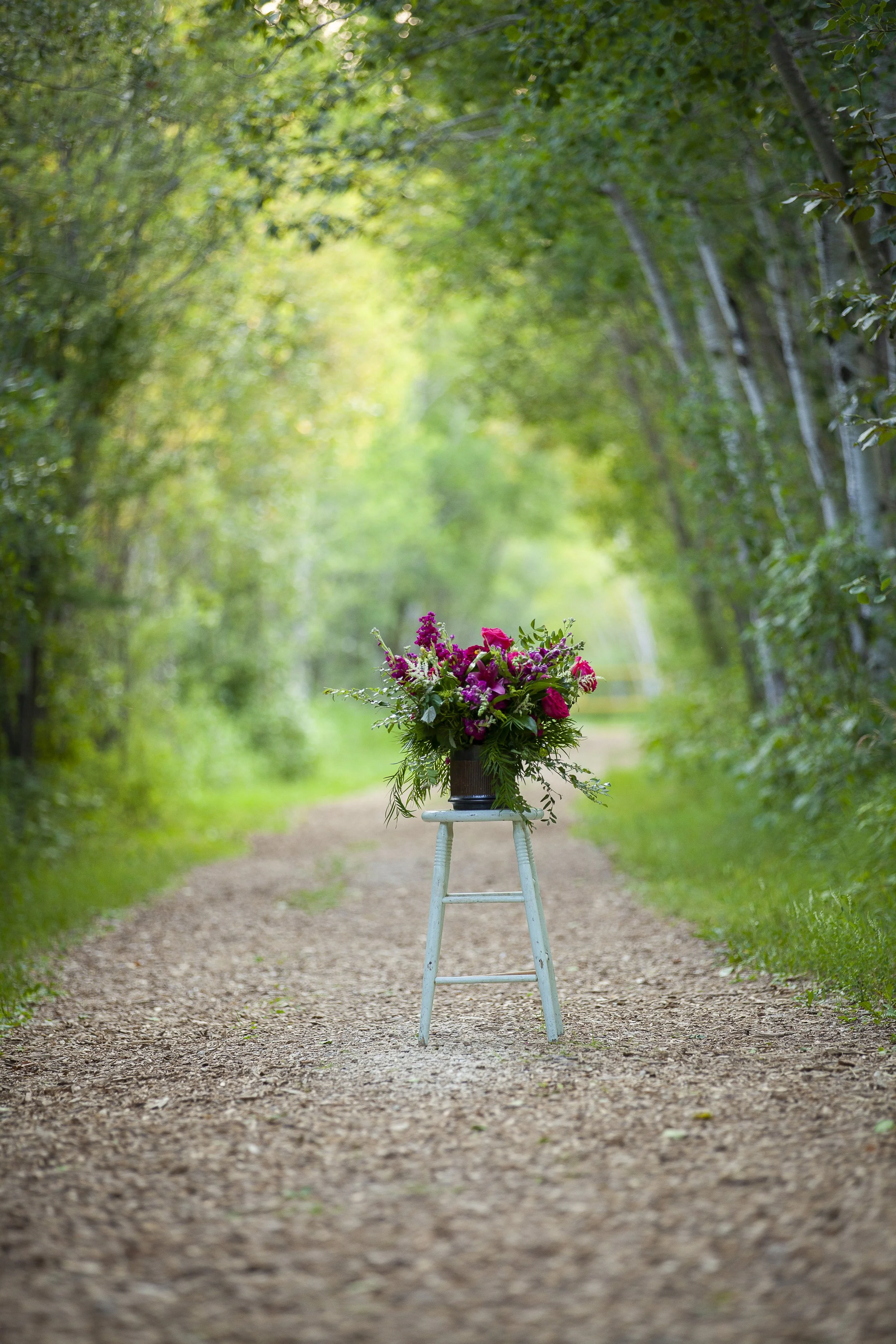 A bouquet of pink and purple flowers on a white wooden stool in a wooded pathway.