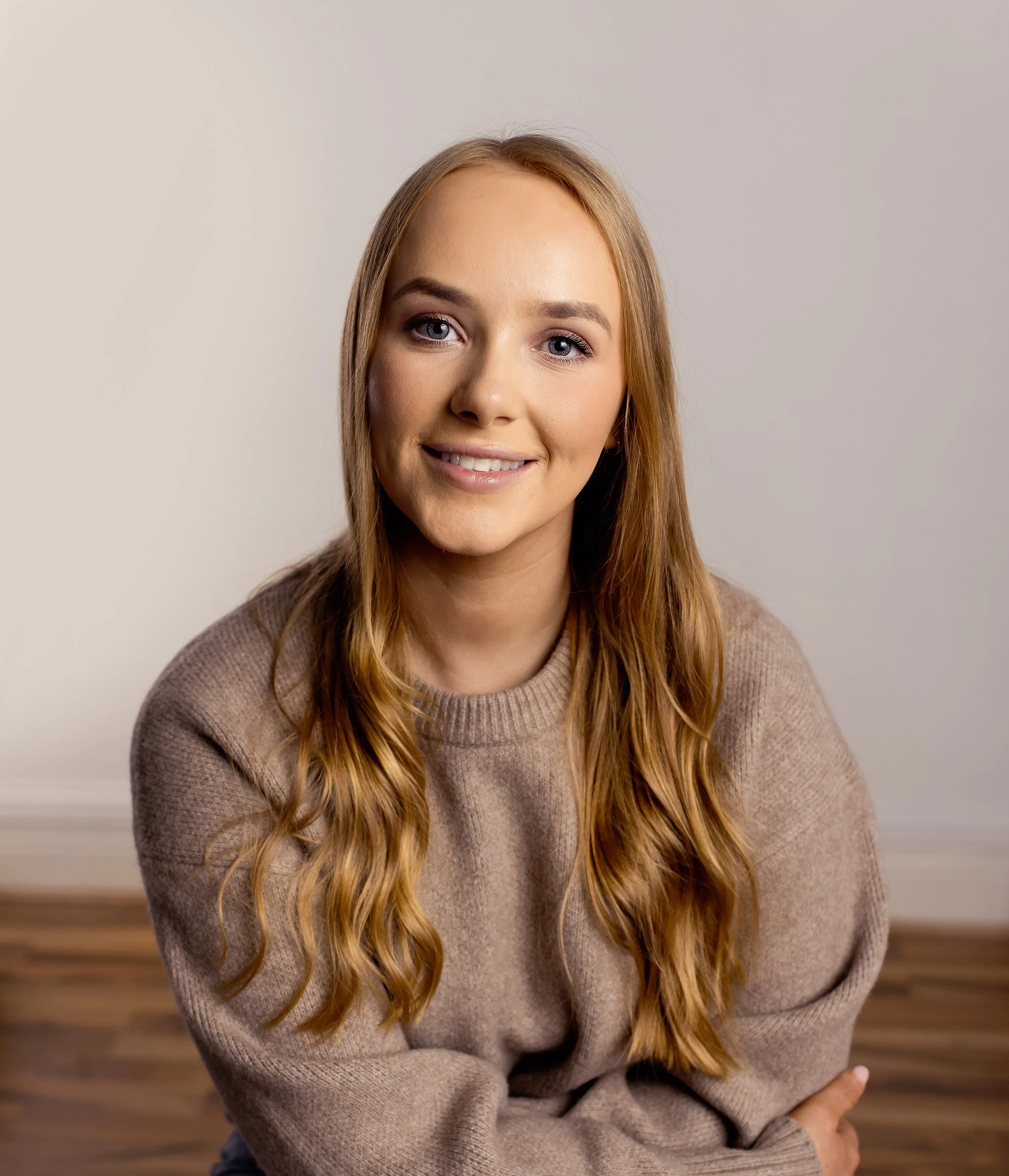A young woman with long, wavy red hair and blue eyes smiling at the camera, wearing a beige sweater, in a room with a plain light-colored wall and wooden floor.