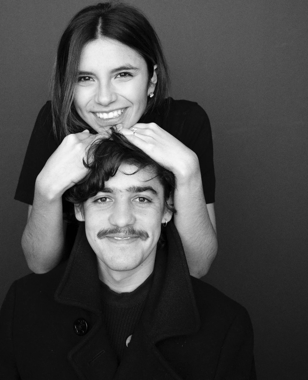 Retrato en blanco y negro de una mujer sonriendo y un hombre con bigote y una sonrisa, ambos miran hacia la cámara. La mujer está detrás de él, descansando sus manos en su cabeza.
