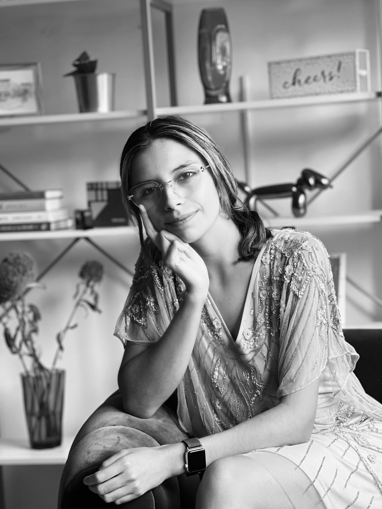 Retrato de una mujer joven con gafas y vestido claro sentado en un sillón, en un ambiente de oficina o estudio, con estanterías y libros en el fondo.