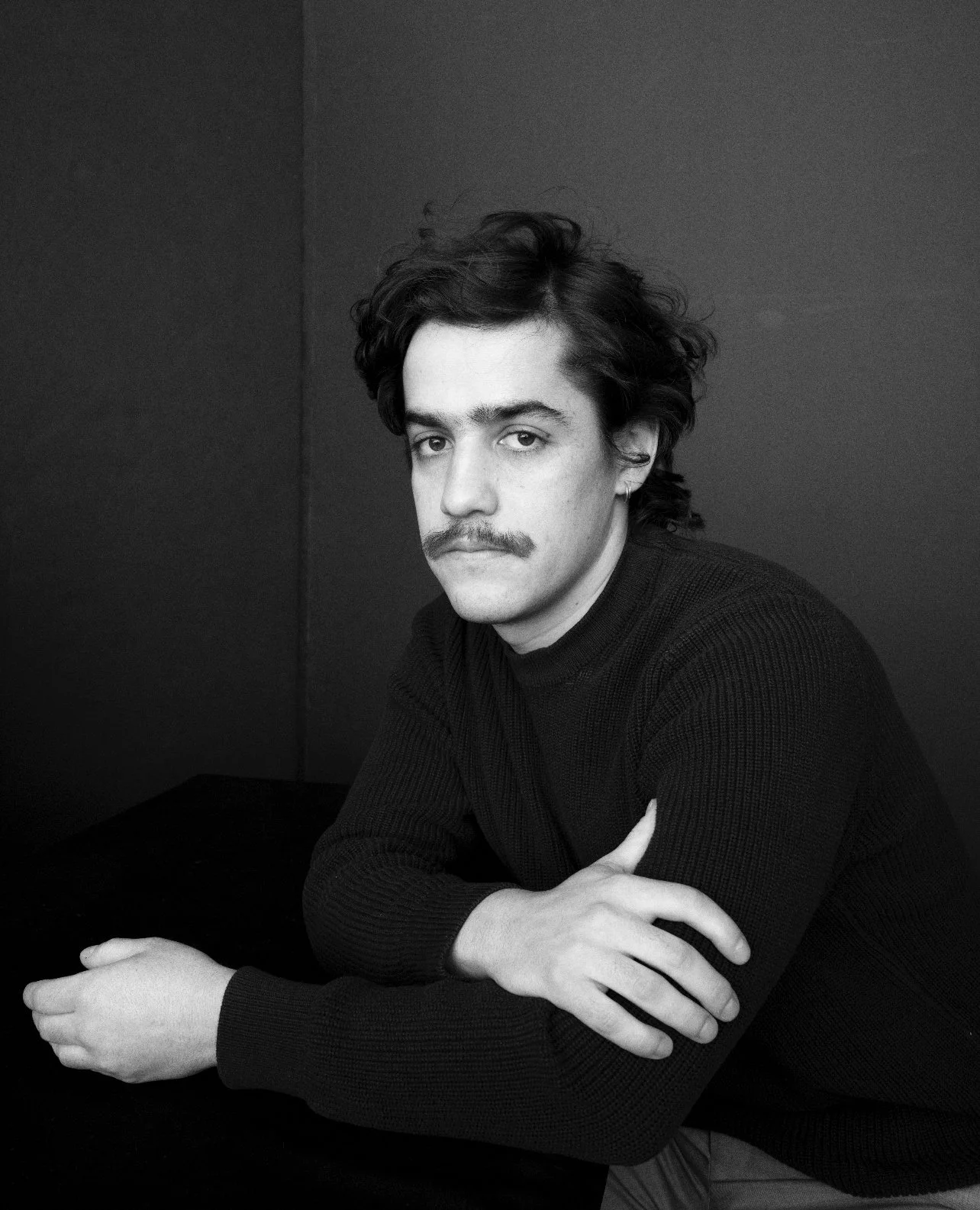 Retrato en blanco y negro de un hombre con cabello rizado y barba, vistiendo un suéter negro, sentado con los brazos cruzados sobre una mesa, mirando hacia la cámara, fondo oscuro.