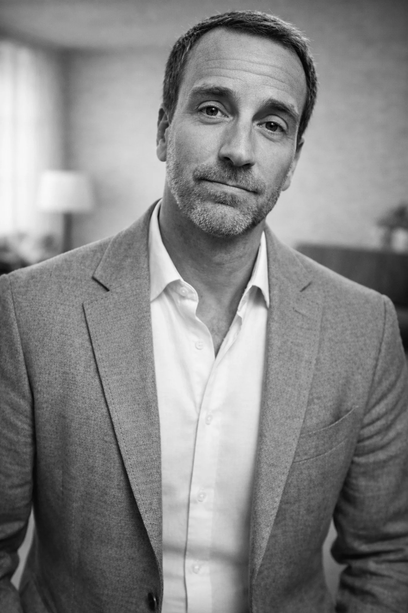 Black and white portrait of a middle-aged man with short hair, slight beard, wearing a light-colored shirt and a textured blazer, looking at the camera with a neutral expression, in a room with a blurred background.