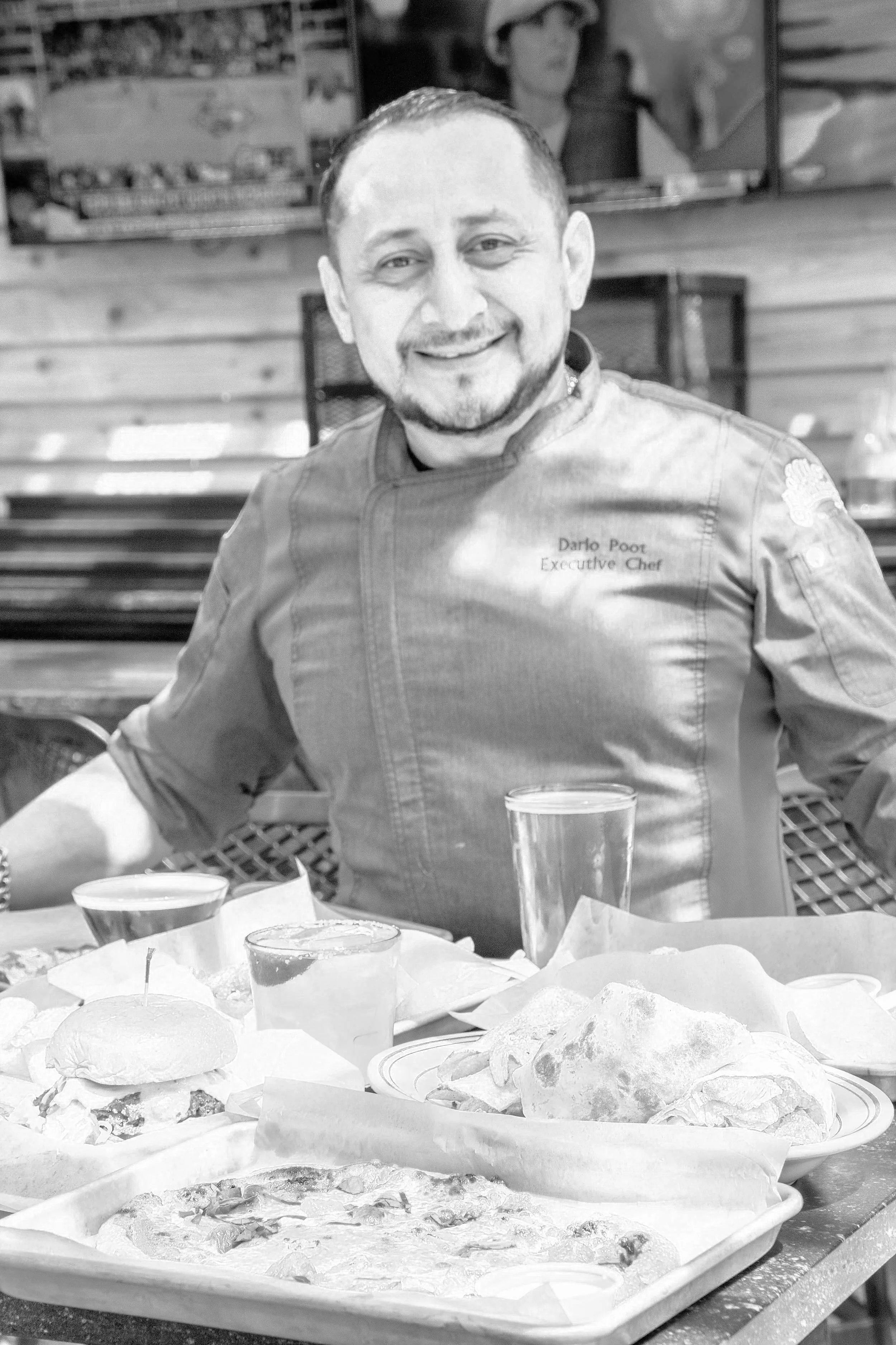A smiling man in a chef's coat sitting at a table with pizza, drinks, and food in front of him, in a restaurant setting.