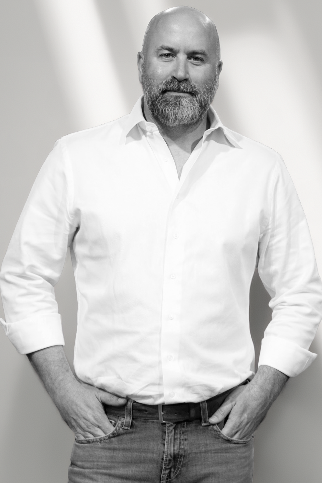 A man with a beard, wearing a white shirt with sleeves rolled up and jeans, stands with hands in pockets, smiling slightly at the camera.
