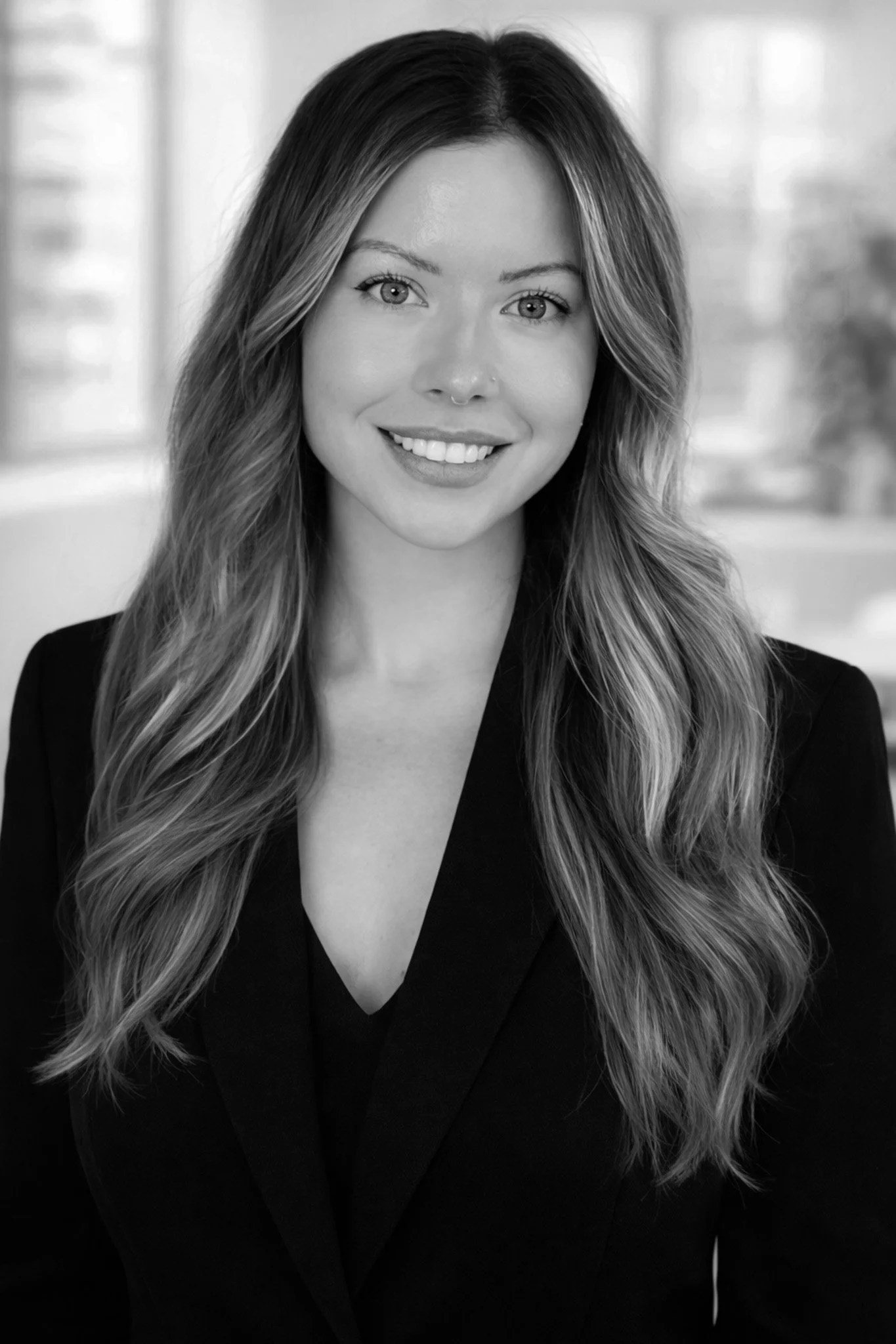 Black and white portrait of a woman with long, wavy hair, smiling, wearing a dark blazer, against a blurred indoor background.