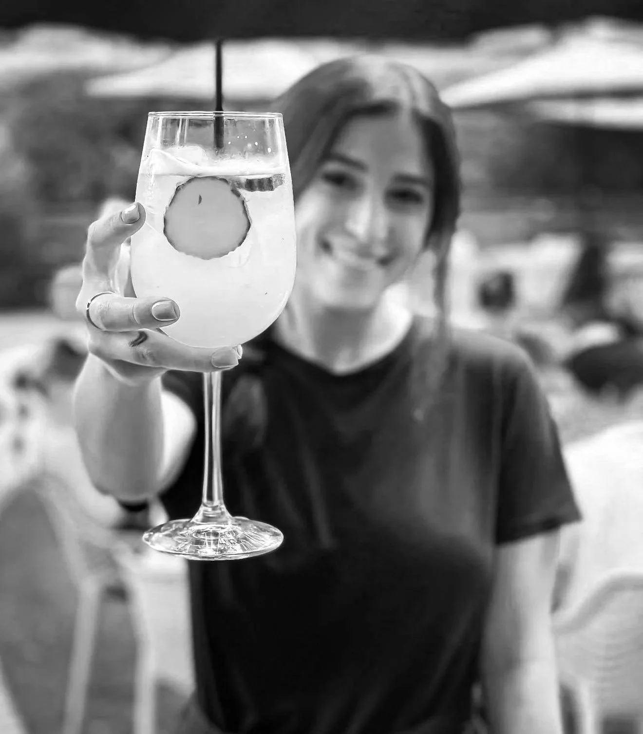 A woman smiling and holding a wine glass with a beverage, ice, and a lemon slice at an outdoor setting.
